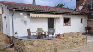 A stone-walled building with a tiled roof, a small patio with a table and chairs, and a window with a potted plant. - Vacation apartment in country house style. small, cozy and quiet location (Wasserlosen)