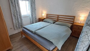A bedroom with a wooden bed, two bedside tables, a window with curtains, and a wall with a textured finish. - Vacation apartment in country house style. small, cozy and quiet location (Wasserlosen)