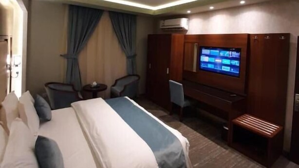 A hotel room with a bed, a desk with a computer, a TV, and an air conditioning unit. - Neroz Inn Furnished Apartments (Jeddah)