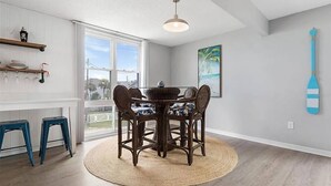 A dining area with a round table, wicker chairs, and a woven rug. There are two blue stools and a hanging light fixture above the table.