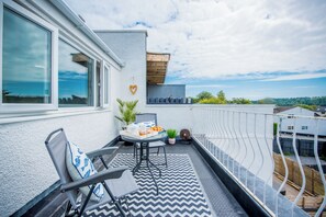 A balcony with a table set for two, a chair, a heart-shaped decoration, and a view of the sea.