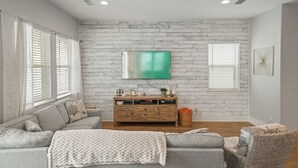 Un salon avec un canapé gris, un meuble TV en bois et un grand téléviseur à écran plat fixé sur un mur en briques blanches.