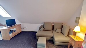 Un espace de vie compact comprenant un canapé, un pouf, un téléviseur posé sur un meuble en bois, une lampe et une petite table de chevet en bois.