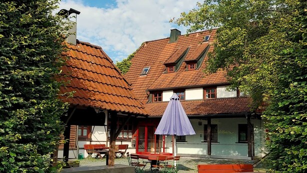 A traditional building with a tiled roof, wooden benches, and a covered seating area with a purple umbrella. - Holiday apartment "Rudolphstein" – 2-4 Guests (Warmensteinach)