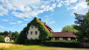 A large, two-story building with a sloped roof, surrounded by greenery and a gravel path. - Holiday apartment "Rudolphstein" – 2-4 Guests (Warmensteinach)