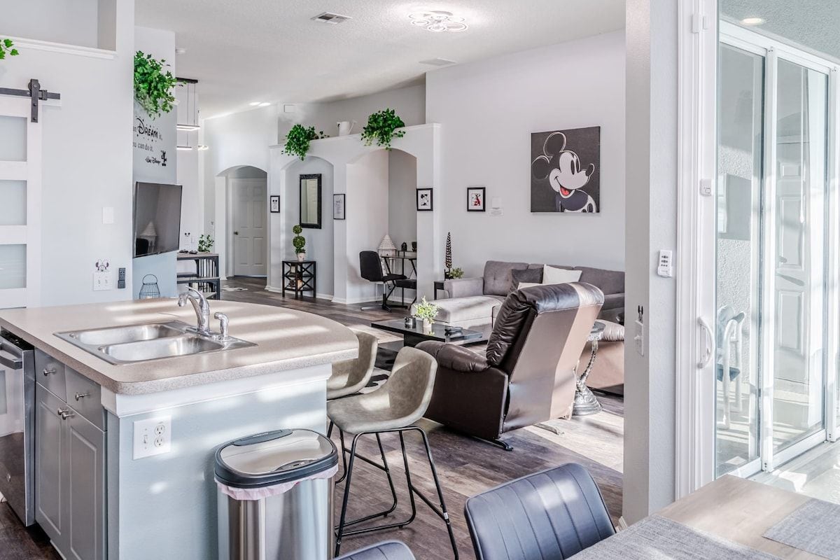 A modern kitchen with a central island, a dining area with a table and chairs, a living room with a sofa and a coffee table, and a glass door leading to an outdoor area.