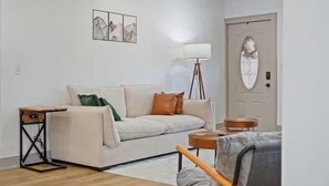 A living room with a sofa, a side table, a lamp, and a door with a glass panel. - Stylish Bungalow. Sleeps 6. Wi-fi. Pets welcome. (Oklahoma City)