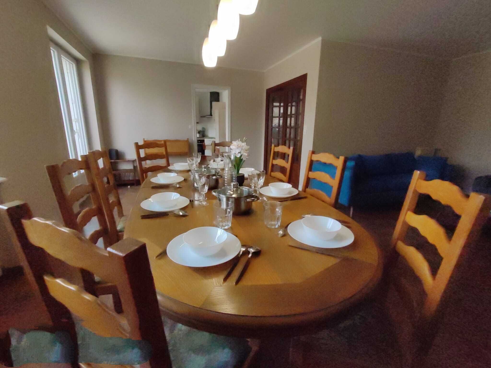 Uma área de refeições com uma mesa de madeira redonda posta para quatro pessoas, rodeada por cadeiras de madeira.
