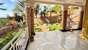 A covered outdoor area with tiled flooring, columns, and a view of a garden with a stone wall and a metal gate. - Mu Rugo by Iwacu Stays (Kigali)
