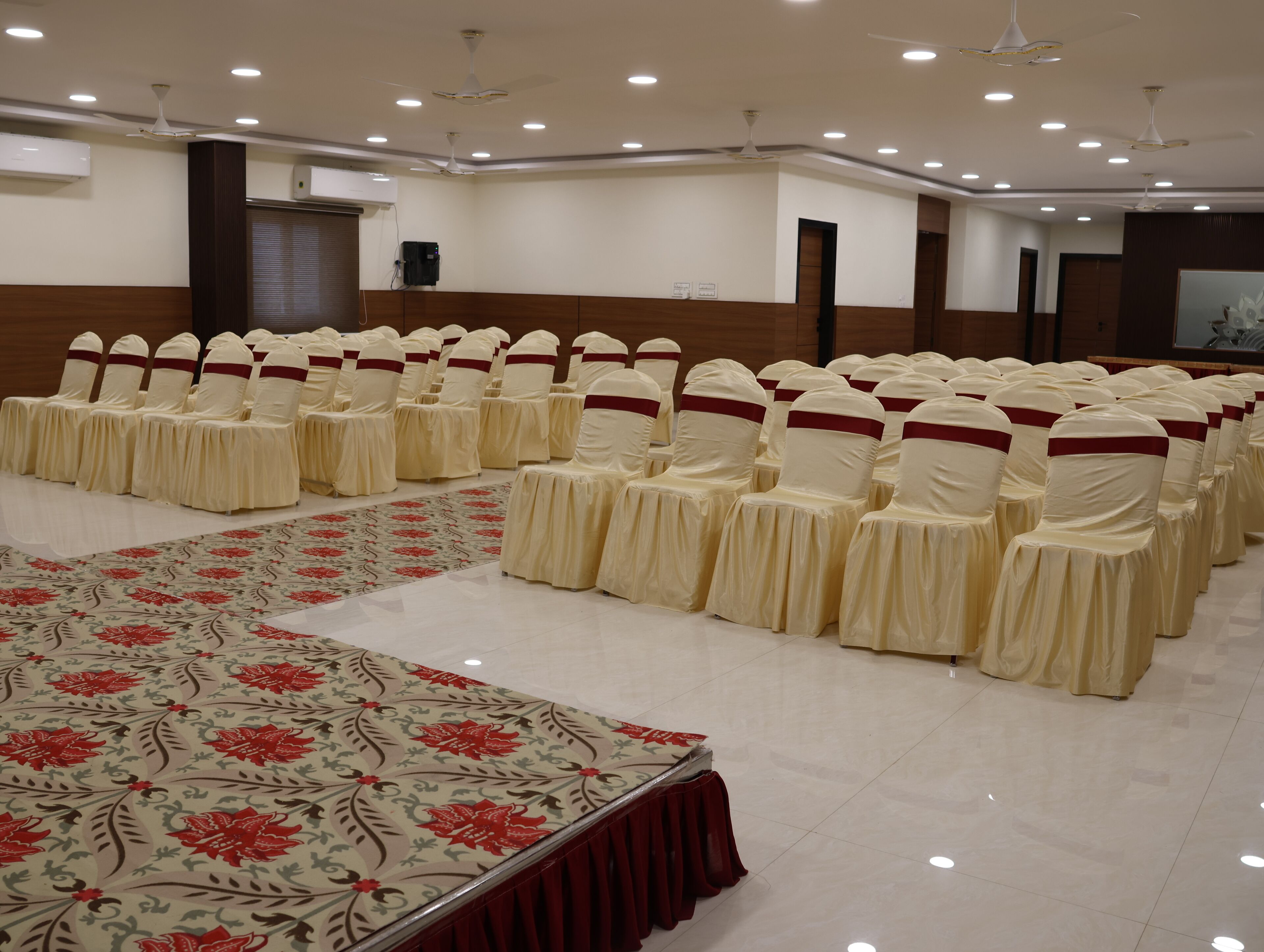 Un salón de conferencias con filas de sillas adornadas con lazos blancos y rojos, un mantel floral grande y ventiladores de techo.