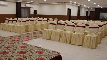 Un salón de conferencias con filas de sillas adornadas con lazos blancos y rojos, un mantel floral grande y ventiladores de techo.