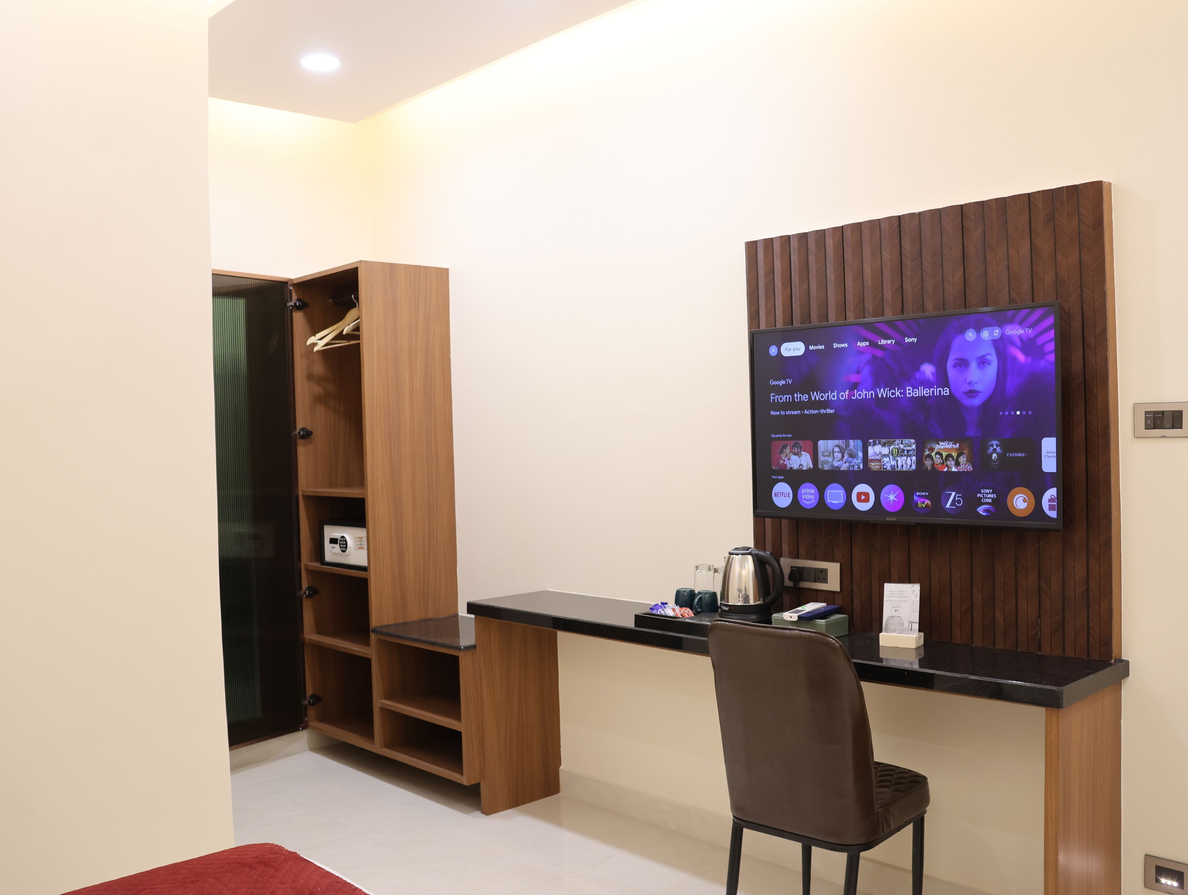 A modern room with a flat-screen TV mounted on the wall, a wooden desk, a chair, and a built-in wardrobe with shelves.