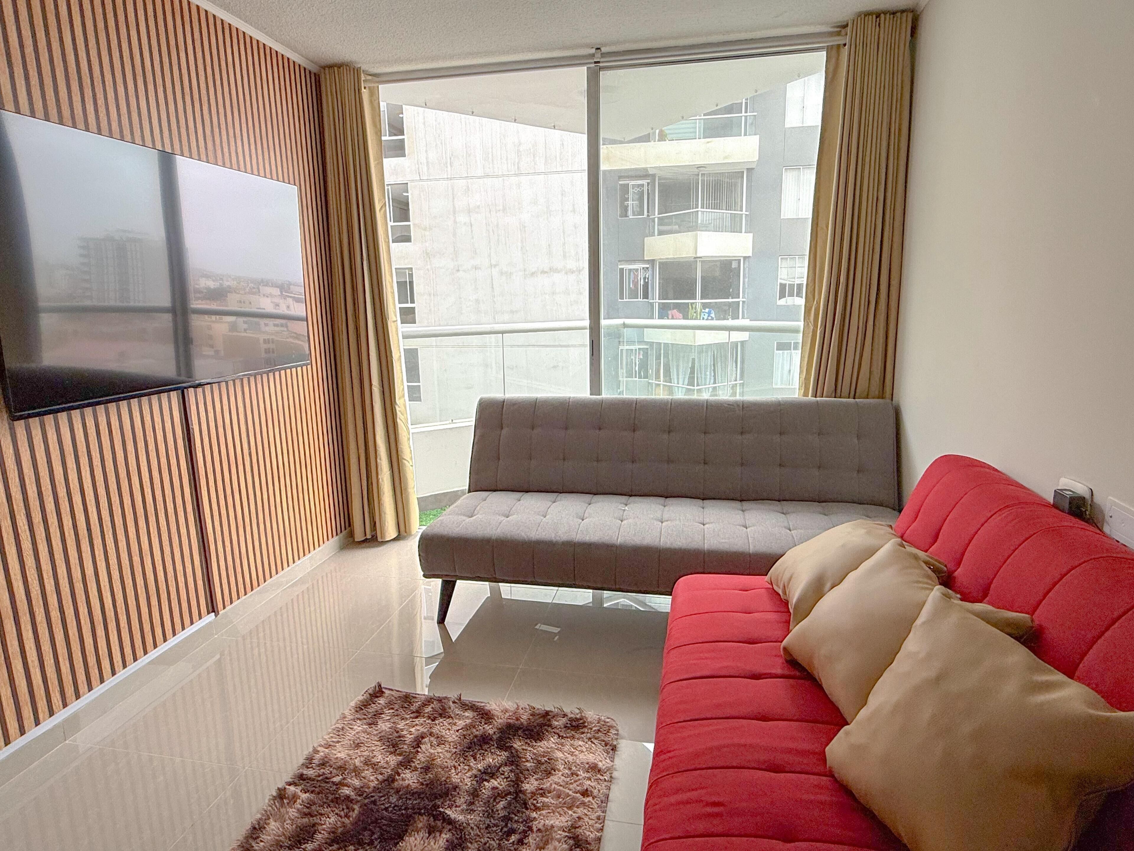 Una sala de estar moderna con un sofá gris, un sofá rojo, un televisor de pantalla plana montado en una pared de paneles de madera y un gran ventanal con cortinas.