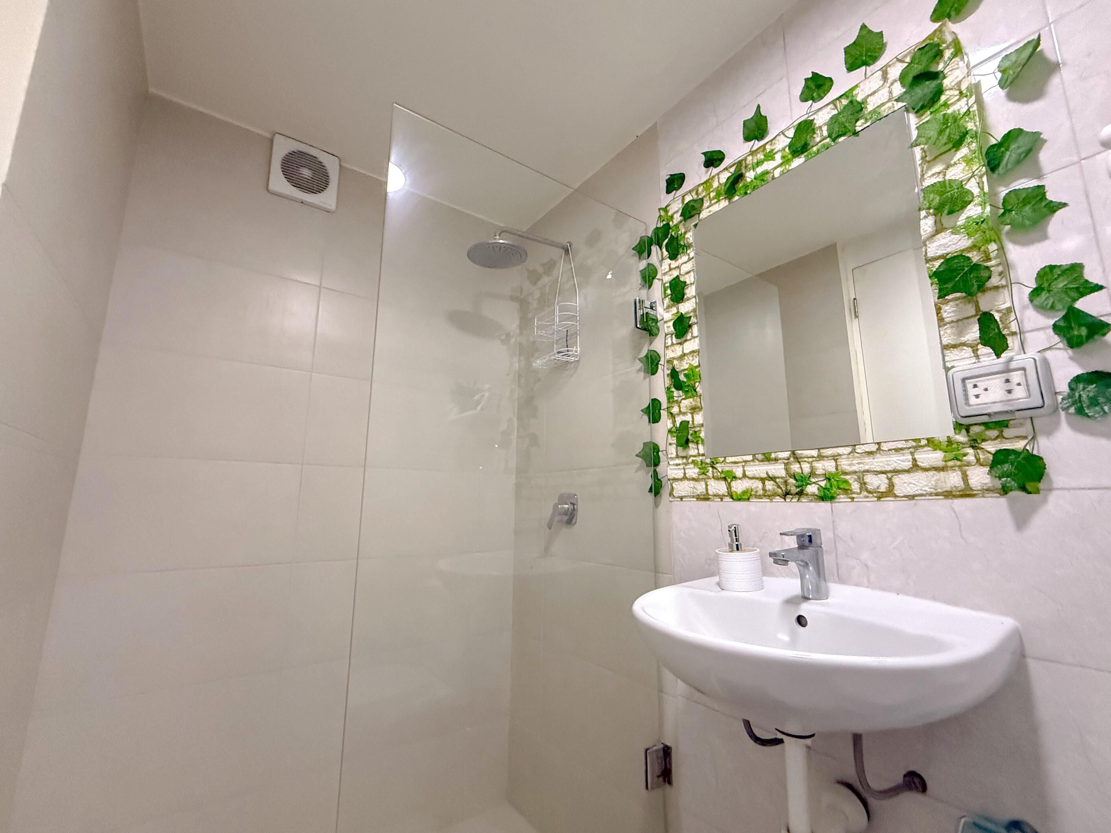 Une salle de bain avec un lavabo blanc, un miroir orné d’une bordure verte en forme de feuilles, et un espace douche.