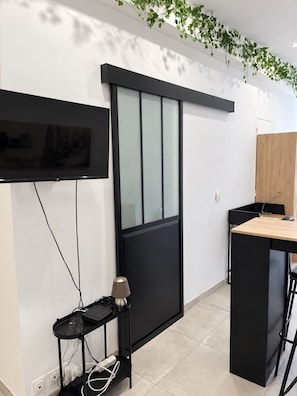 A modern interior with a flat-screen TV mounted on the wall, a sliding door, a small table with a lamp, and a wooden desk. - Renovated studio in the heart of the village, 7 minutes by tram from Montpellier (Saint-Jean-de-Védas)