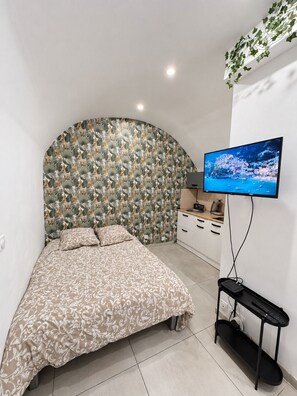 Een slaapkamer met een gebogen hoofdeinde, een bed met een bedsprei met dessin, een aan de muur bevestigde televisie en een klein kitchenettegedeelte.