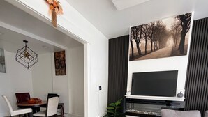 Una sala de estar moderna con un televisor de pantalla plana, un sofá gris, una zona de comedor con una mesa y sillas de madera, y un pasillo que conduce a otra habitación.