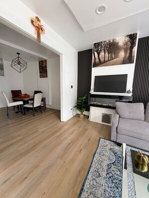 Una sala de estar moderna con un televisor de pantalla plana, un sofá gris, una zona de comedor con una mesa y sillas de madera, y un pasillo que conduce a otra habitación.