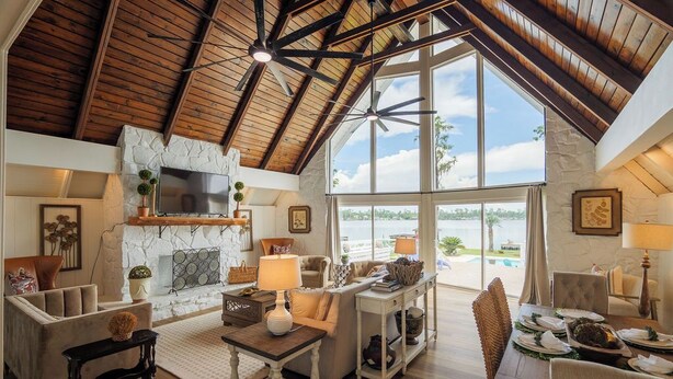 A spacious living room with a high ceiling, stone fireplace, large windows, and a dining area with a table set for four.
