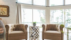 A room with two beige armchairs, a round side table with a mirror mosaic, and a view of trees through large windows.