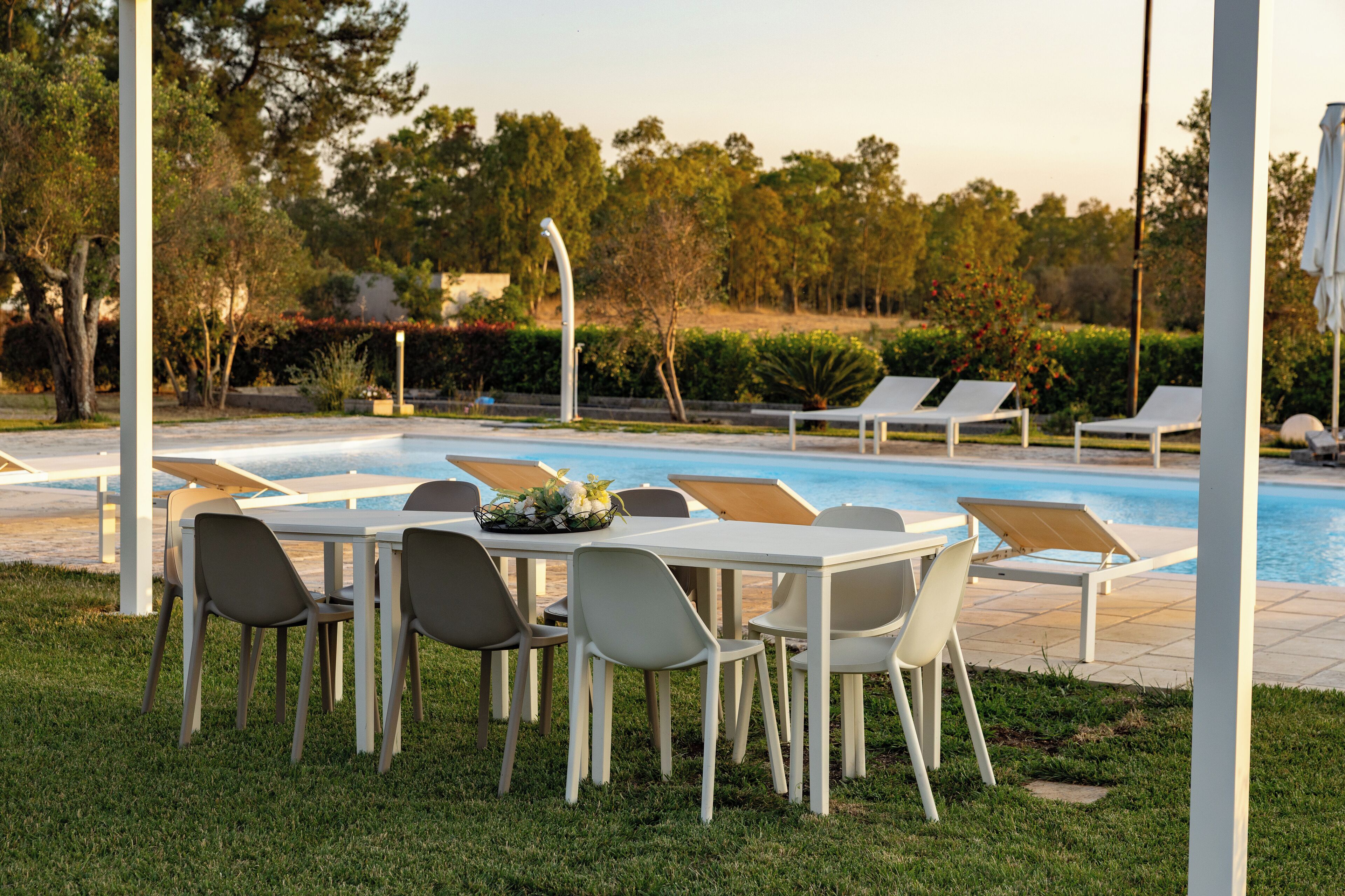 Un área junto a la piscina con sillas de descanso y una mesa con sillas, rodeada de follaje y árboles.