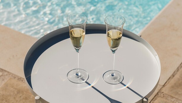 Taula rodona amb dos gots de vi blanc, situada al costat d'una piscina d'aigua cristal·lina.