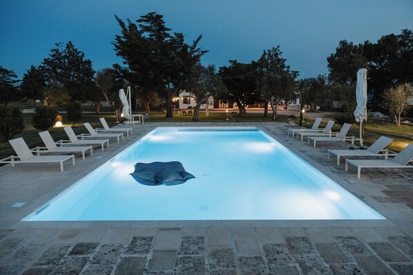 Une piscine extérieure dotée de chaises longues et de parasols, entourée d’arbres et avec un bâtiment en arrière-plan.