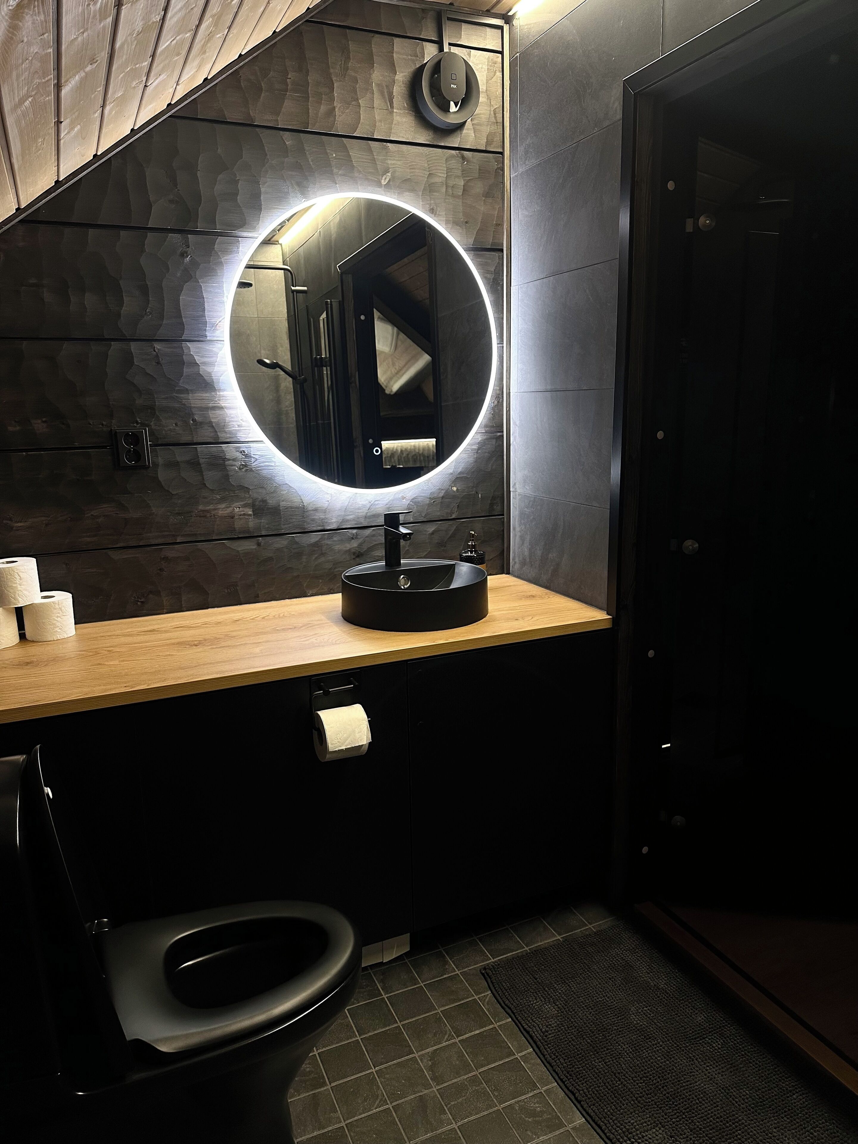A modern bathroom with a wooden vanity, a round mirror with a light, a toilet, and a dark cabinet.
