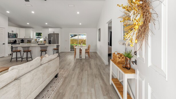 A modern kitchen with white cabinets, a dining area with a wooden table and chairs, and a living room with a white sofa.