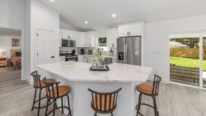 A modern kitchen with white cabinets, a large island with bar stools, stainless steel appliances, and a view of a backyard.