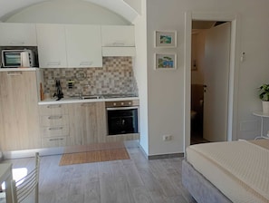 A compact kitchen with a microwave, oven, and sink, a small dining area with a chair, and a bedroom with a bed.
