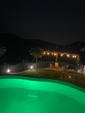 A nighttime view of a pool area with a green-lit pool, a seating area with a table and chairs, and a railing.