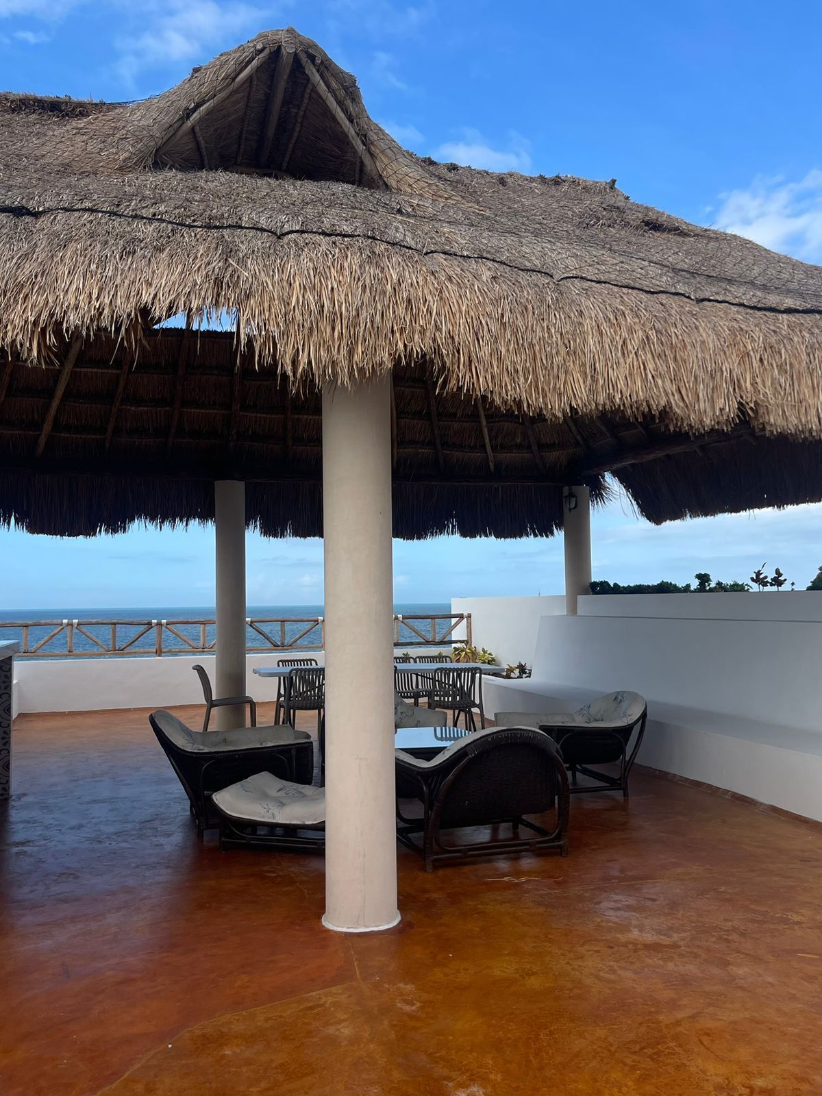 An outdoor seating area with a thatched roof, tables, and chairs, overlooking the sea.