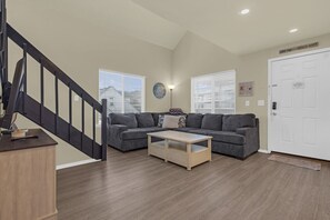 Un salon avec un canapé gris, une table basse en bois, un escalier et une porte blanche avec une pancarte.