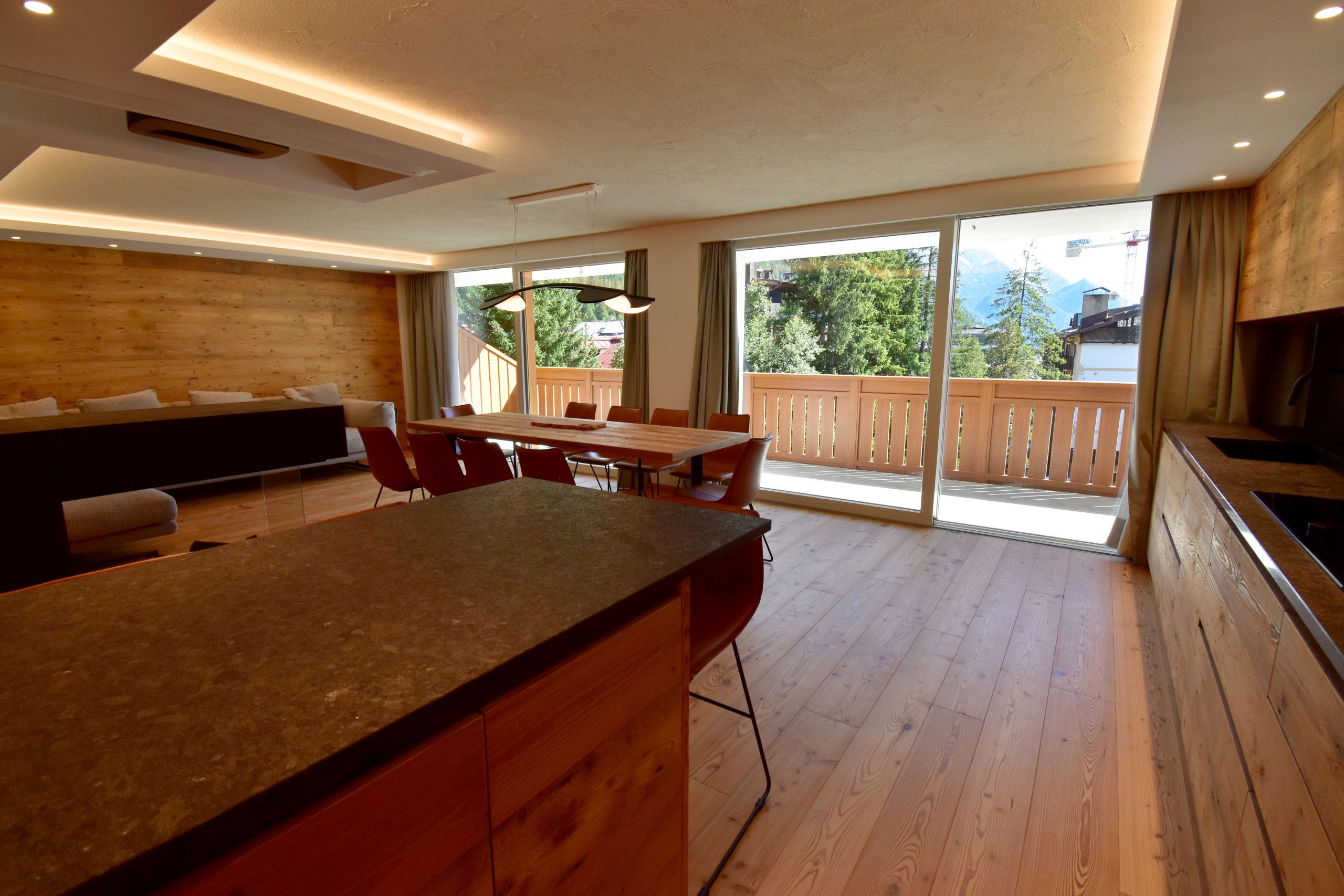 Een moderne keuken met een groot schiereiland, een eethoek met stoelen, en uitzicht op een houten terras en bergen door grote schuifdeuren.
