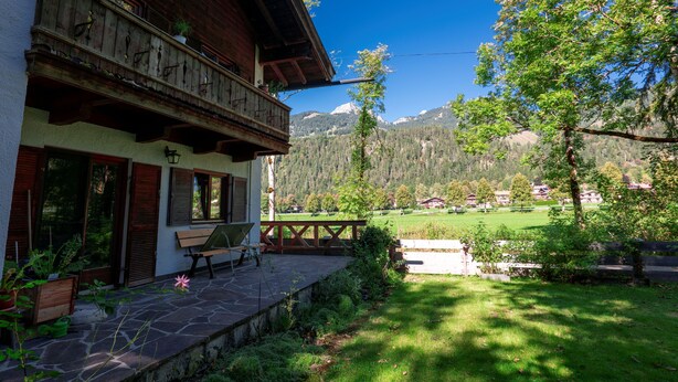 A wooden building with a balcony, a stone patio, and a view of a green field and mountains. - Haus Luise - Cozy vacation apartment with mountain views (Bayrischzell)