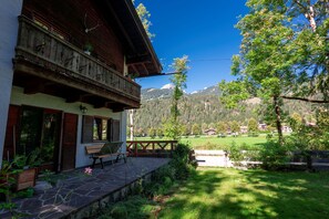 A wooden building with a balcony, a stone patio, and a view of a green field and mountains. - Haus Luise - Cozy vacation apartment with mountain views (Bayrischzell)