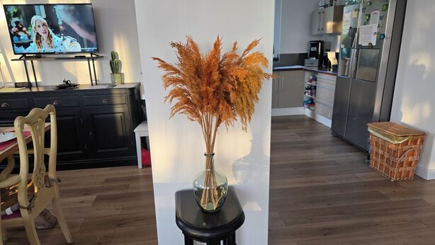 A modern kitchen with a dining area, a flat-screen TV, and a vase with dried plants on a stool.
