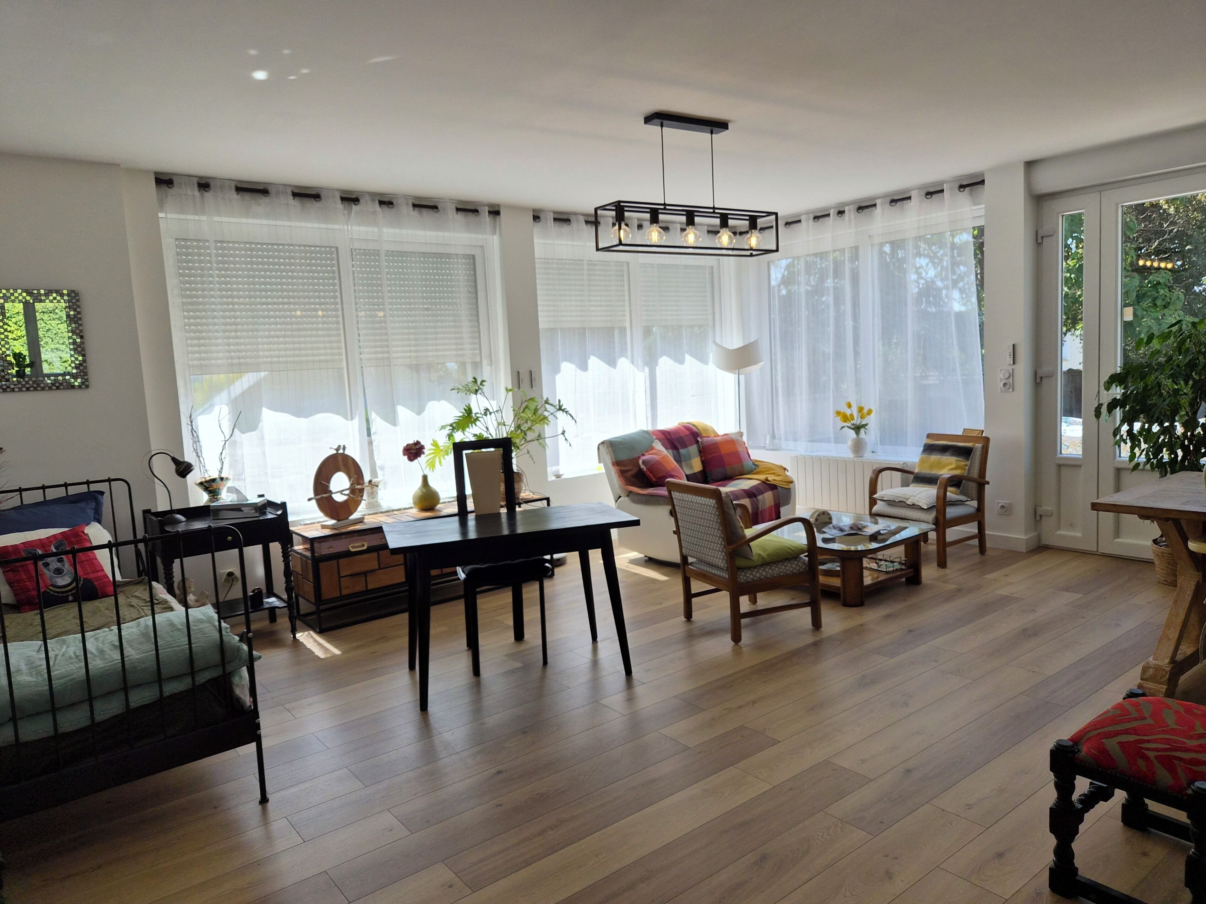 A well-lit living room with a wooden floor, a dining table, and a sofa. There are large windows with blinds, a ceiling light fixture, and a potted plant.