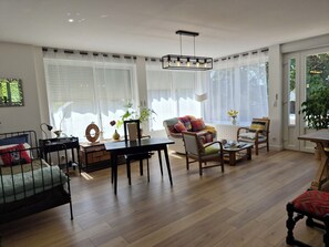 A well-lit living room with a wooden floor, a dining table, and a sofa. There are large windows with blinds, a ceiling light fixture, and a potted plant. - A beautiful and very spacious two-bedroom apartment in Bretteville-sur-Odon, in the Venoix neighborhood of Caen. (Bretteville-sur-Odon)