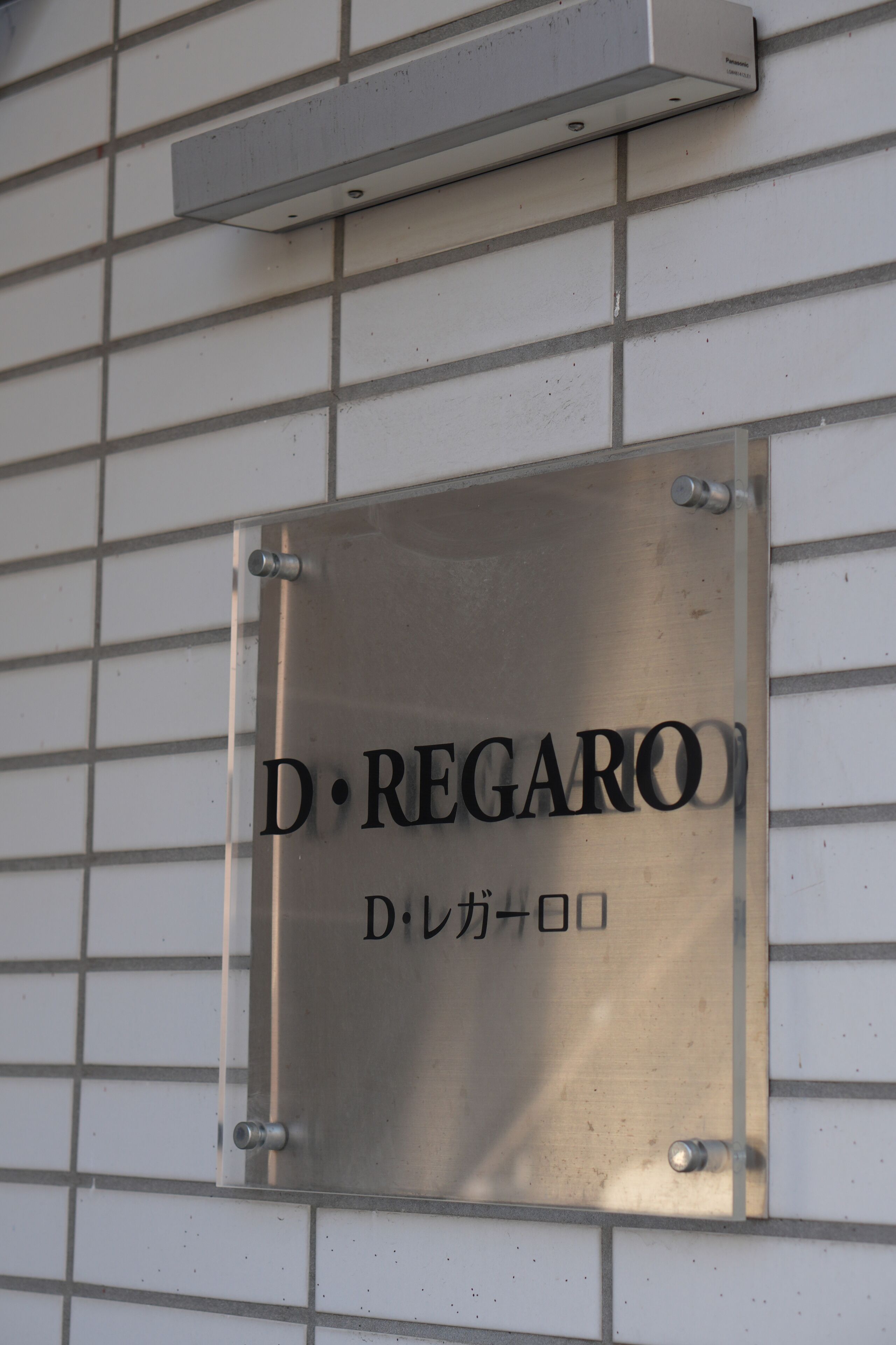 Una placa metálica con el texto "D. Regar" y caracteres japoneses sobre una pared de azulejos.