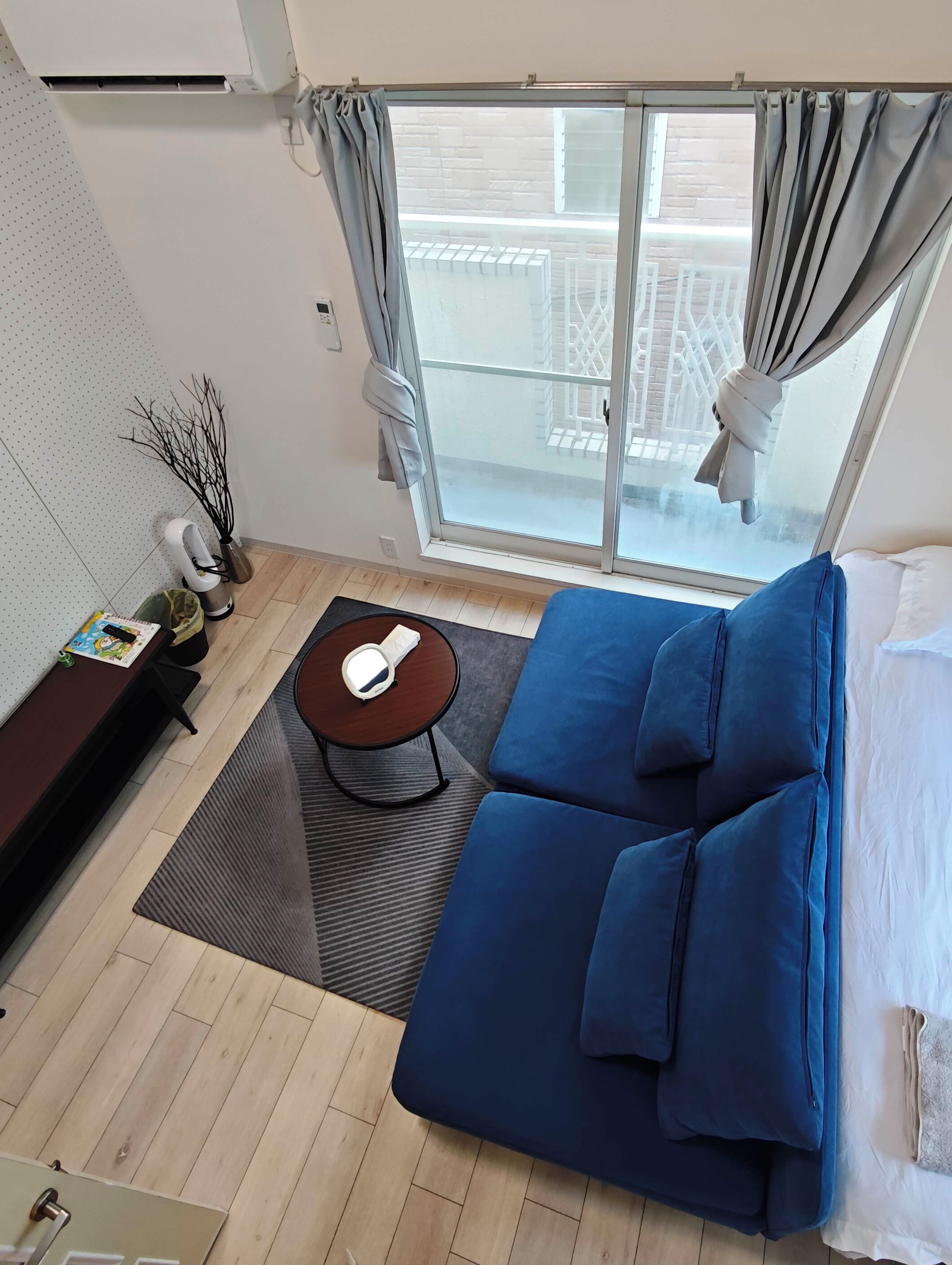 Una habitación con un sofá azul, una mesita con un objeto blanco, una ventana con cortinas y vistas a un balcón.