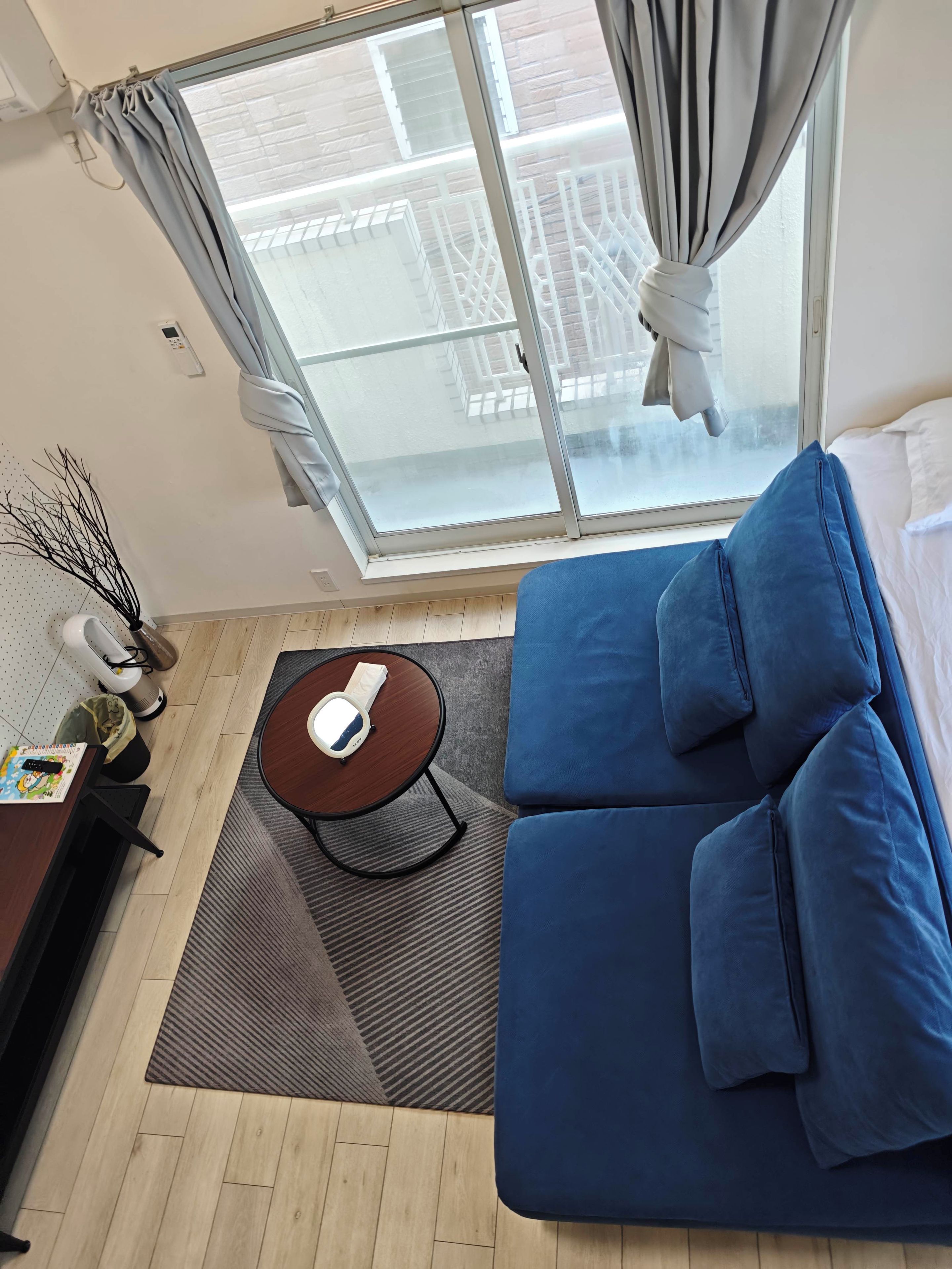 Quarto com sofá azul, mesa redonda pequena com um objeto branco, janela com cortinas e vista para um edifício lá fora.