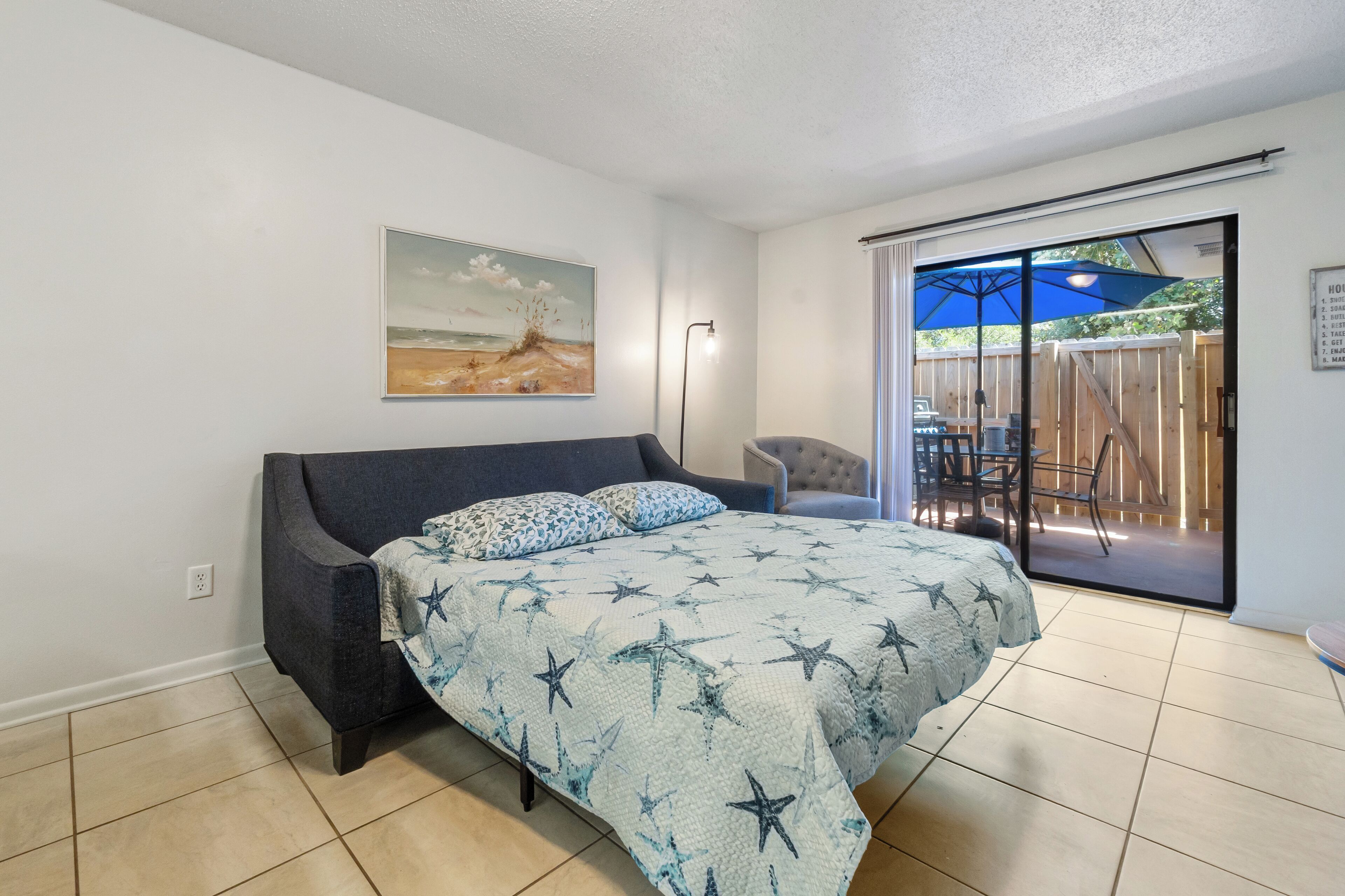 A bedroom with a bed, a painting, a chair, and a view of an outdoor area with a wooden deck and an umbrella.