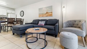 Un salon moderne avec un canapé d’angle, un fauteuil gris, une table basse ronde et un tableau représentant une scène de plage.