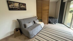 A modern living room with a grey sofa, a patterned throw pillow, a small side table, a woven basket, and a large window with a view of greenery. - Apartment Cannes garden 5mn close to downtown 5mm Gare plages Croisette (Cannes)