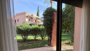 A view through a window with a view of a courtyard, a building with a pink facade, and a brick pathway. - 2-bed Apartment with Garden & shared Pool- Moroccan - Modern Style (Marrakech)