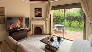 A living room with a fireplace, a television, and a view of a garden through a glass door. - 2-bed Apartment with Garden & shared Pool- Moroccan - Modern Style (Marrakech)