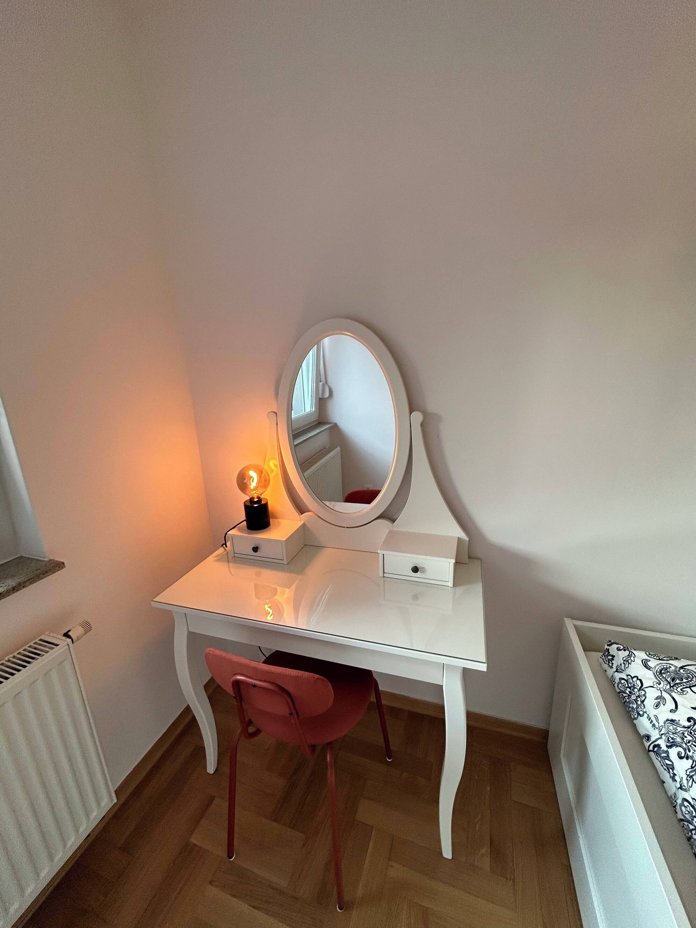 Et hvidt toiletbord med et rundt spejl, en lille lampe og en rød stol.