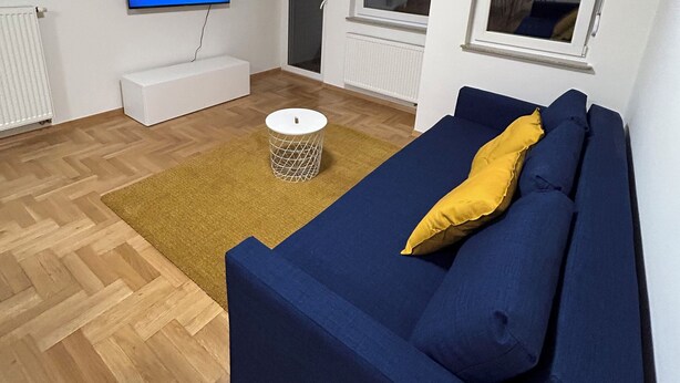 A modern living room with a blue sofa, a yellow pillow, a round white side table, and a flat-screen TV mounted on the wall. - Apartment Fran (Zagreb)